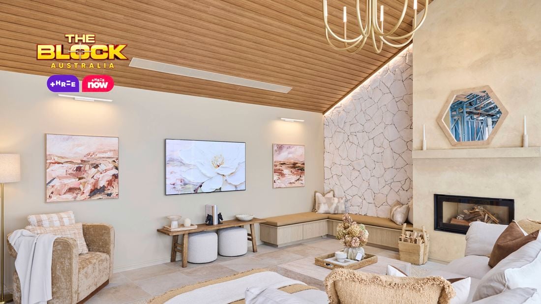 A modern living room with a high wooden ceiling and a large chandelier. The space includes a white and beige sectional sofa with pillows, an armchair draped with a blanket, and two small round stools. A fireplace on the right is framed by stacked firewood, candles, and an octagonal mirror above. The back wall features two paintings flanking a central TV displaying flowers. The left wall is clad in textured stone.