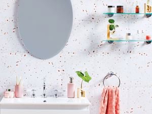 A light-coloured bathroom with speckled tiling, a basin with light pink accessories, a salmon coloured hand towel, an oval mirror and two floating glass shelves in a decluttered bathroom.