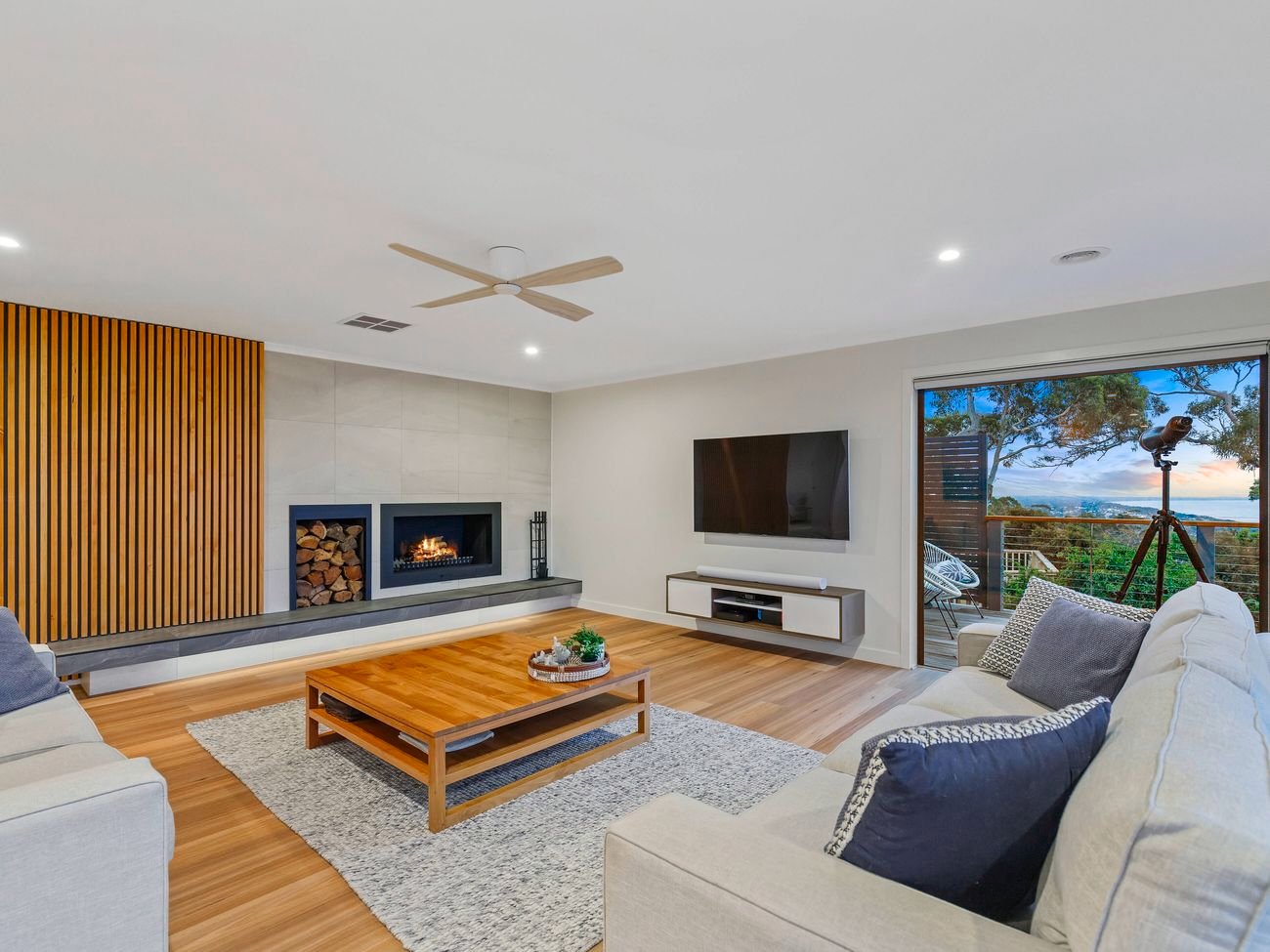 Modern living room with fireplace, wooden paneling, scenic window view, light-colored sofas, blue cushions, coffee table, and wall-mounted TV.