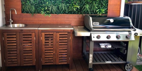 Outdoor decking area with a BBQ, bench and sink in front of a timber panelled wall with artificial plants