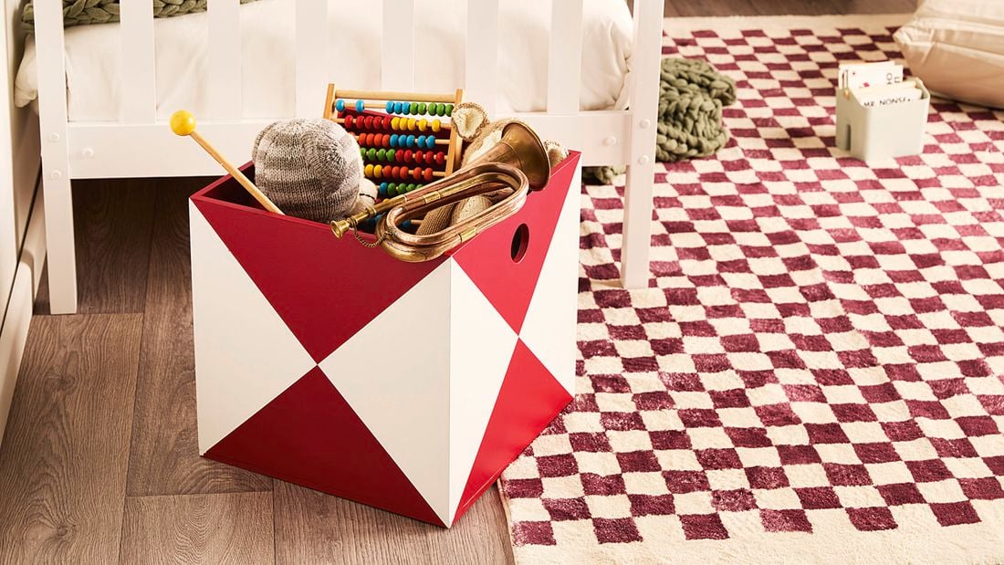 Red-and-white geometric storage box in a child’s room holding toys on a burgundy-and-cream chequered rug beside a white bed.