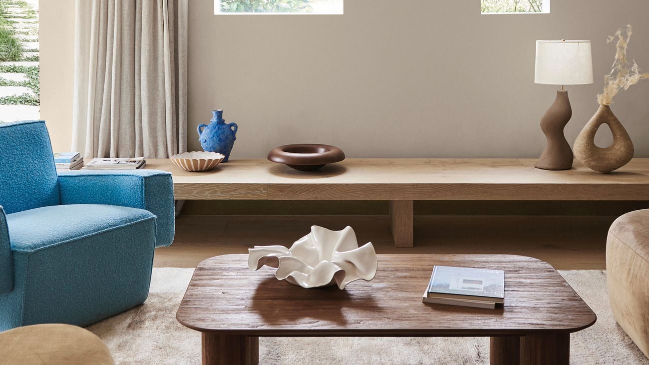 Stylish living room with wooden coffee table, blue armchair and brown couch surrounding it, lamp on long wooden cupboard in background.