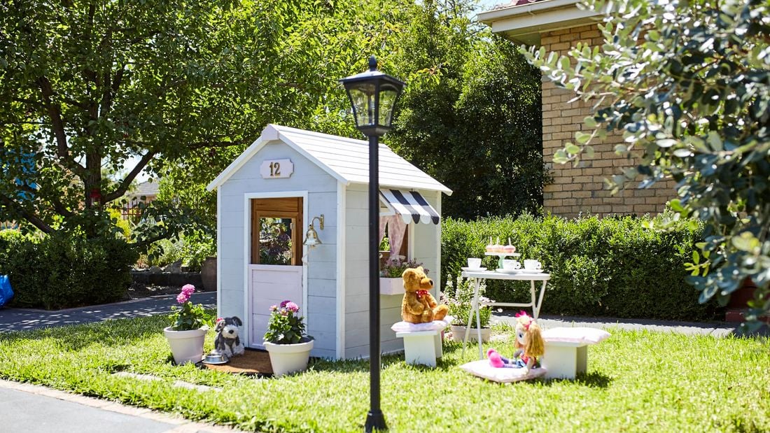 A painted and decorated cubby house with potted plants, a teddy, a doll and a tea set