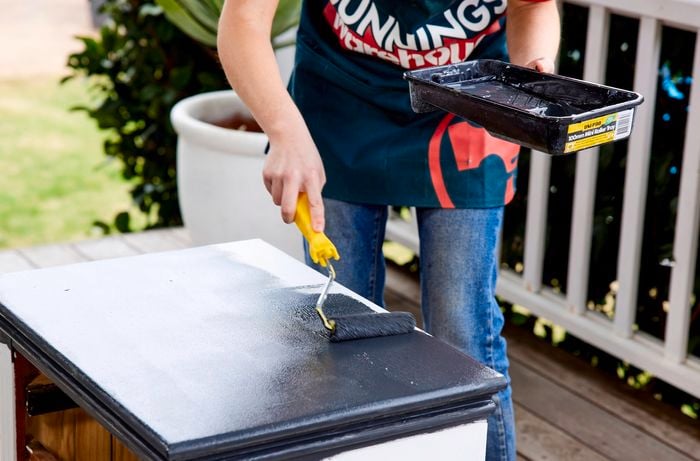 Person applying chalk paint to bedside table with a mini roller while holding tray of paint.