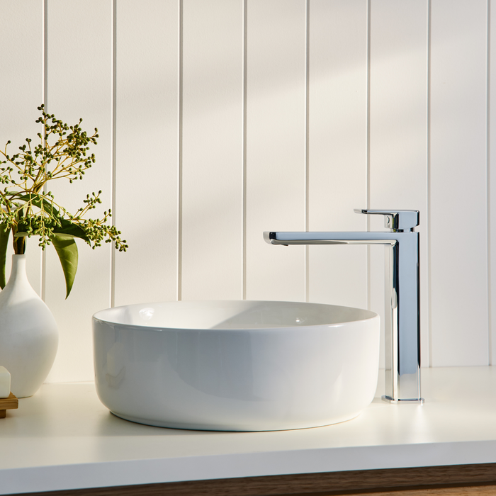 A basin with tap above it, a vase with flowers in it on a white bench top in a bathroom.