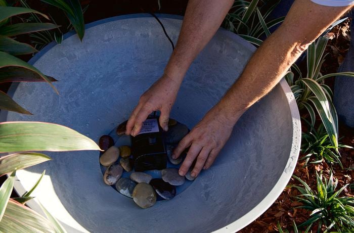 Hands position a water pump surrounded by stones in a large grey bowl.