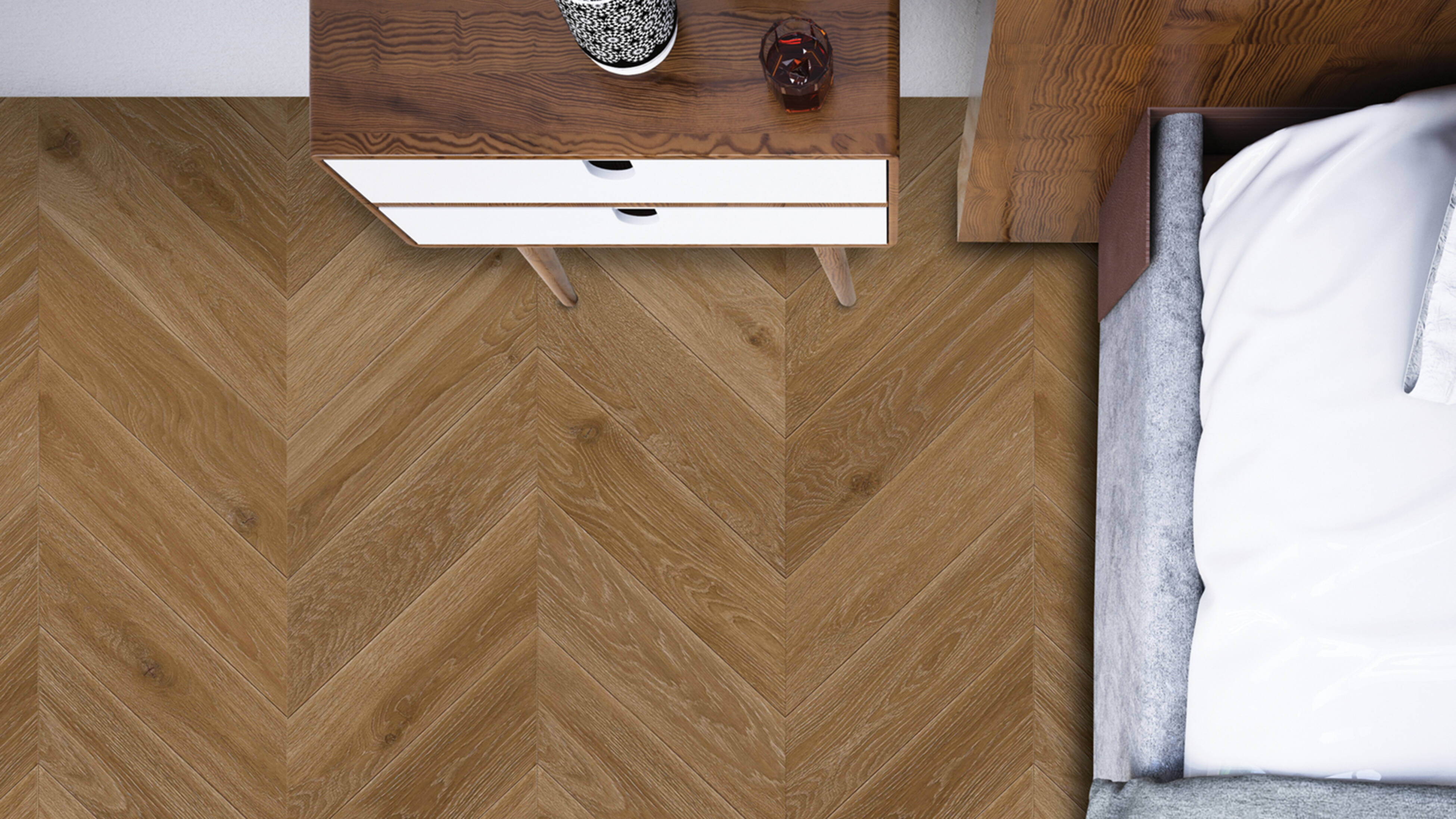  Overhead view of a bedroom with herringbone wooden flooring, a wooden bedside table with white drawers, and part of a bed with white linen.
