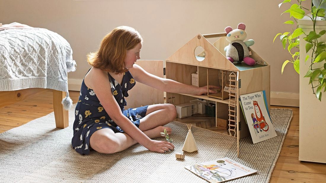 Young girl plays with a wooden dolls house in her bedroom, with books and a soft toy.