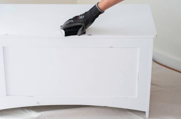 A person is lightly sanding a white timber toy box to prepare the surface for painting, an essential step in creating a DIY games storage box.