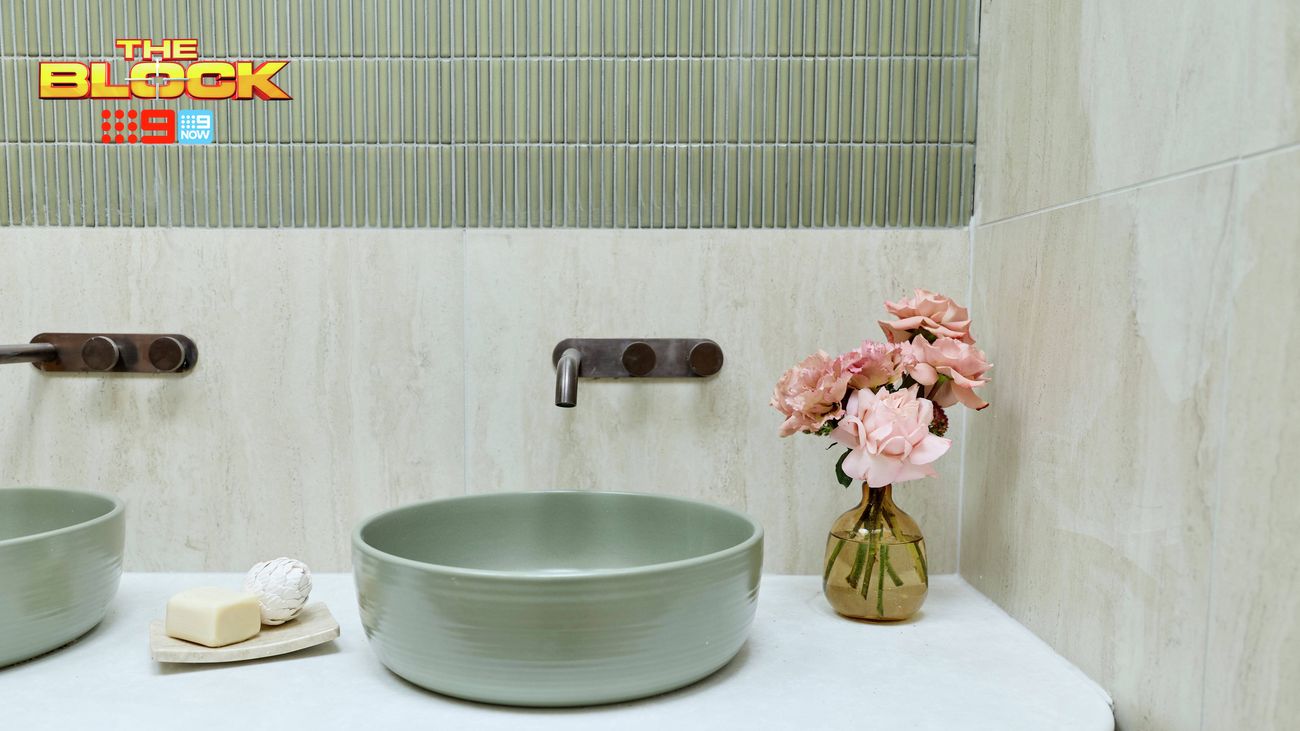 Bathroom vanity with sage green vessel basins, wall-mounted tapware, timber cabinetry, green feature tiles and a vase of pink flowers. “The Block” logo appears in the upper-left corner.