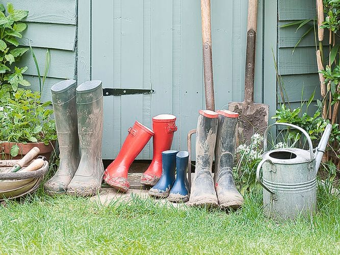 Gardening tools and equipment next to a shed, including gumboots, a shovel, a pitchfork and a watering can.