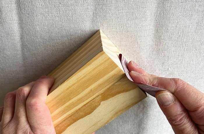  A hand holding a timber block and another hand using sandpaper to sand the side of the block, on a drop sheet.]