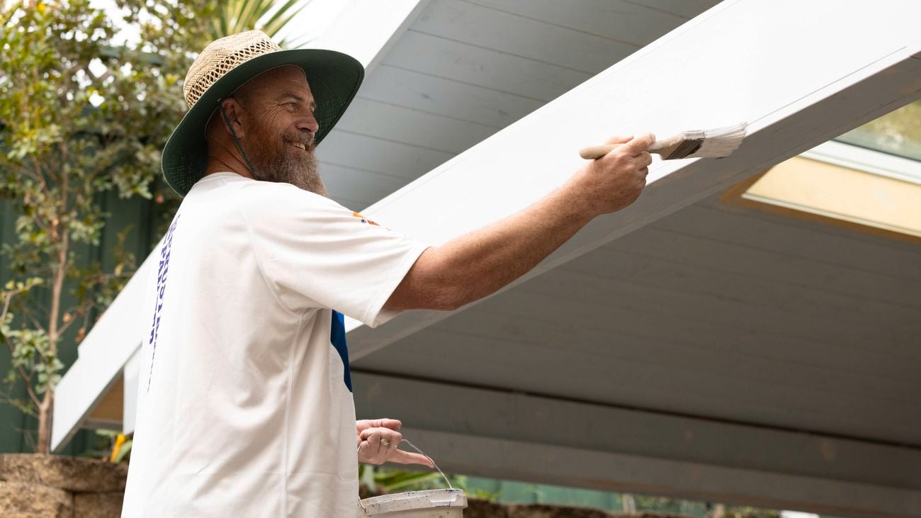 A painter in a wide-brim Bunnings Warehouse straw hat holds a paint bucket while painting the exterior of a house using white paint.]