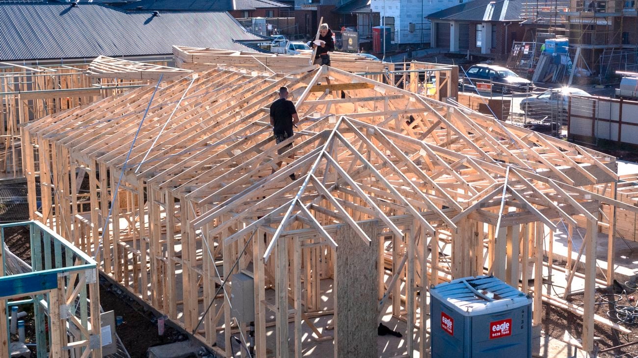Two tradies stand in frame and truss work on a construction site with a fence around it and signage