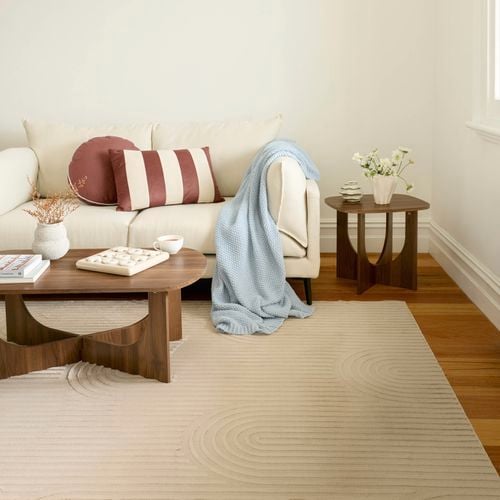 A minimalist living room with a light-coloured sofa, wooden coffee tables, decorative cushions, a blue throw blanket, and natural light from a nearby window.