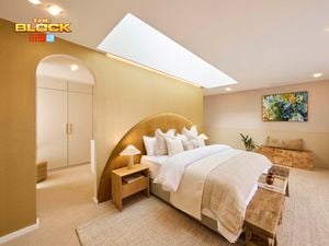 A tastefully decorated bedroom with a large bed centered under a skylight, dressed in a beige blanket and multiple pillows. Two bedside tables with lamps flank the bed. A fireplace is visible on the right, with a bench at the foot of the bed. A colorful painting hangs above a small plant and a piece of furniture. The room features neutral-toned walls and carpeted flooring.