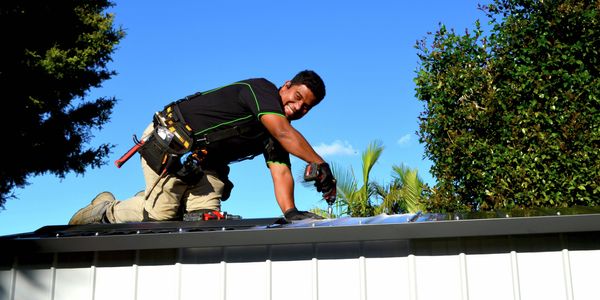 Person kneeling on a metal roof and using a cordless drill to install roofing materials outdoors, with trees and a clear blue sky in the background.