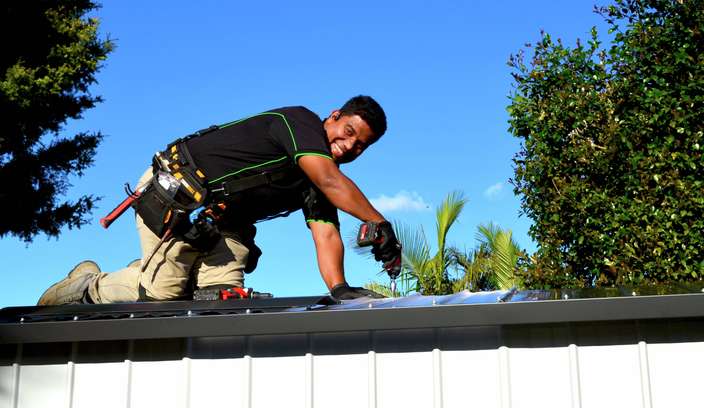 Person kneeling on a metal roof and using a cordless drill to install roofing materials outdoors, with trees and a clear blue sky in the background.
