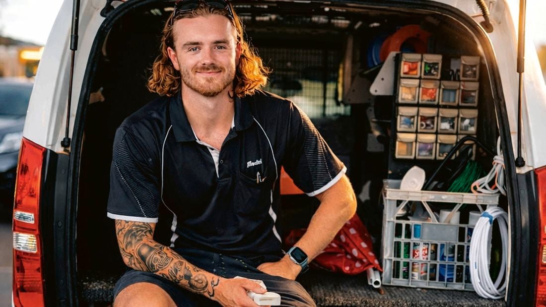 A smiling tradesperson with long hair, a beard, and tattooed arms sits in the back of a work van filled with tools.]