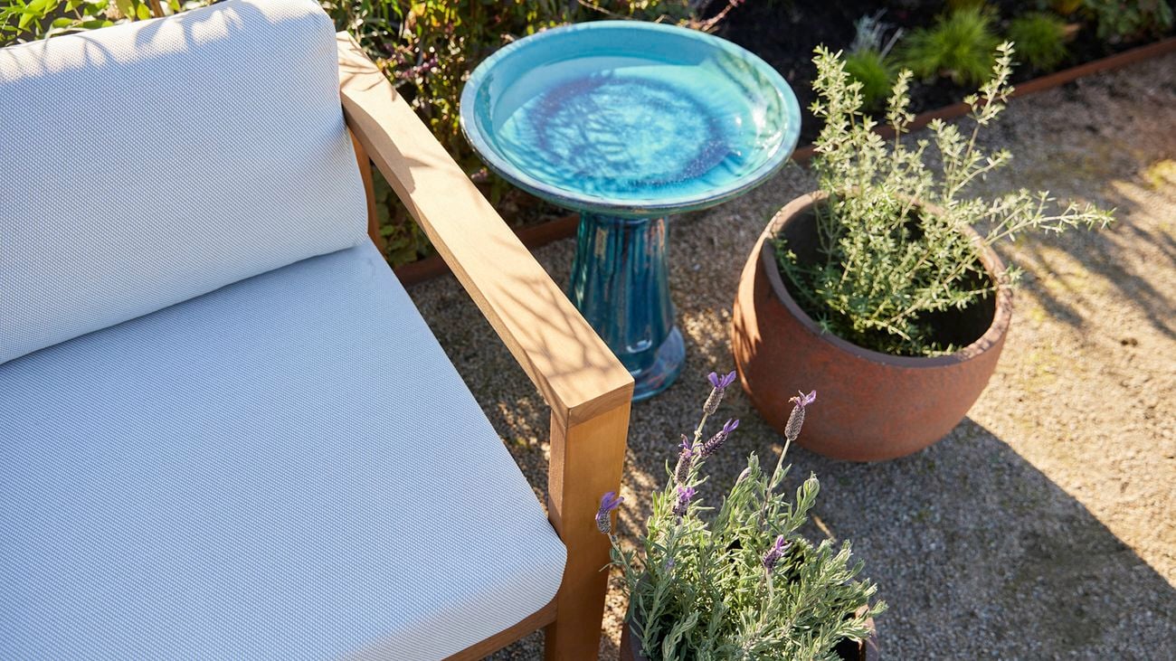 A turquoise bird bath sits on gravel next to a white outdoor bench seat and large pots of lavender and rosemary