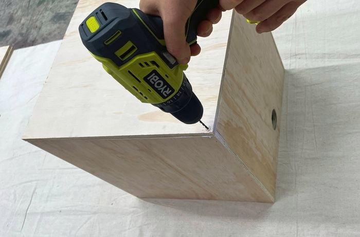 Hands using a cordless drill to screw two plywood panels together at a right angle.