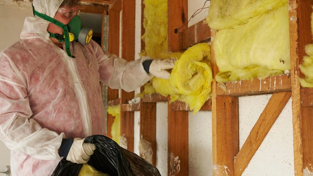 A person wearing protective gear removing insulation batts from a stud wall frame
