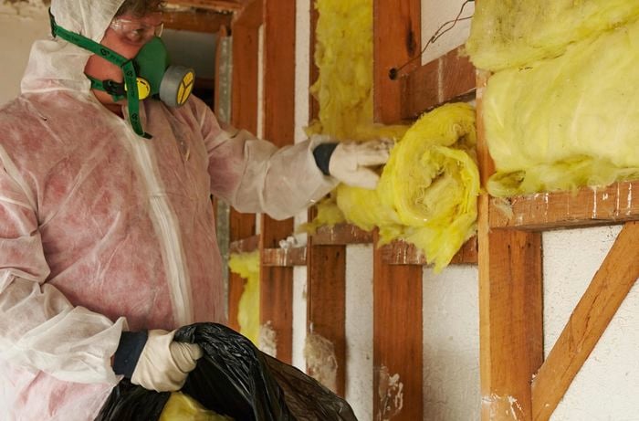 A person wearing protective gear removing insulation batts from a stud wall frame