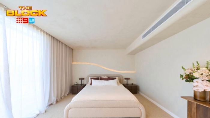 Bright, minimalist bedroom with a cream upholstered bed, floor-to-ceiling sheer curtains, neutral walls, bedside tables, and a vase of flowers. “The Block” logo appears in the upper-left corner.