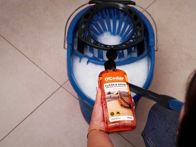 A person holding a bottle of O'Cedar Clean & Shine over a blue bucket. 