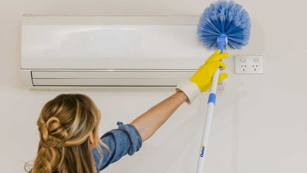 A woman using a cobweb broom on an air conditioning unit.