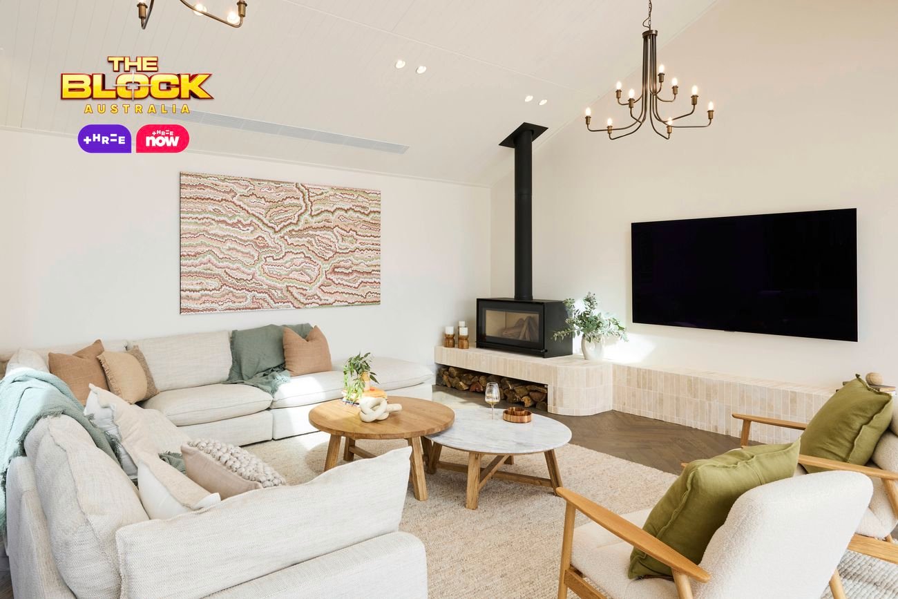 A modern living room with high ceilings, featuring two chandeliers, a wall-mounted flat-screen TV, and a fireplace with logs stored below. The space includes a light-colored sectional sofa with cushions and throws, two armchairs with green cushions, and two round wooden coffee tables. Abstract artwork hangs above the sofa, and large windows bring in natural light. Decorative plants are placed throughout. 