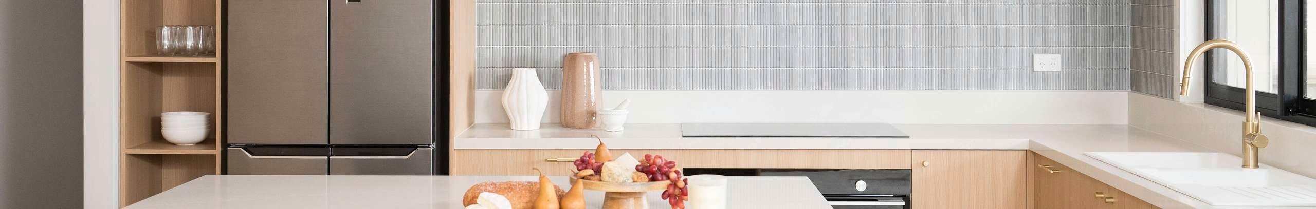 Light brown modern kitchen, with white benchtops with crockery on it, brown cupboards and a fridge in it.