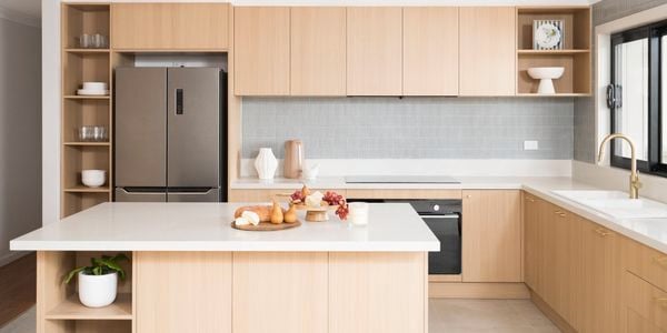 Light brown modern kitchen, with white benchtops with crockery on it, brown cupboards and a fridge in it.