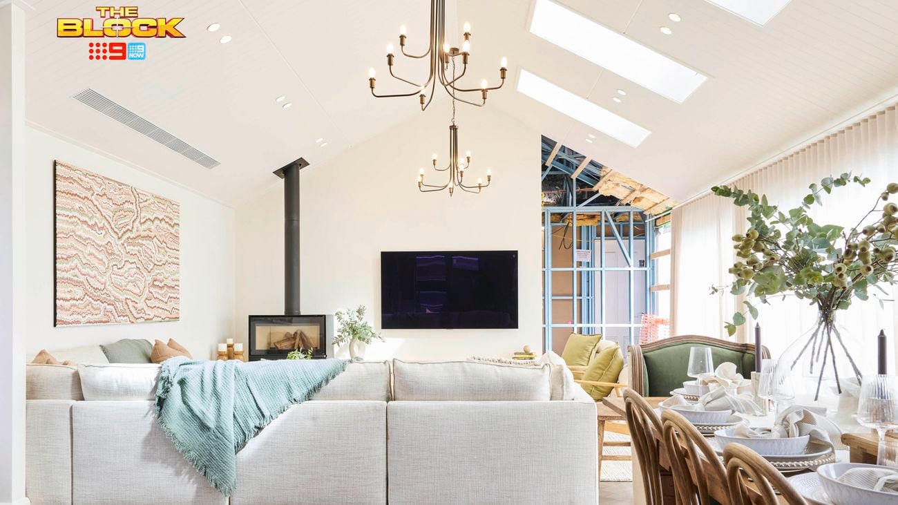 A modern living room with high ceilings and skylights, featuring a large sectional sofa with a green throw blanket facing a wall-mounted TV and fireplace. A chandelier hangs overhead. To the right, a dining table set for six with white plates and napkins sits near large windows with sheer curtains. Greenery in vases decorates the table. 