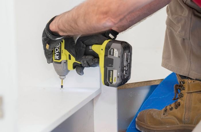 A person wearing gloves and work boots is using a power drill to unscrew a base cabinet during a kitchen renovation.