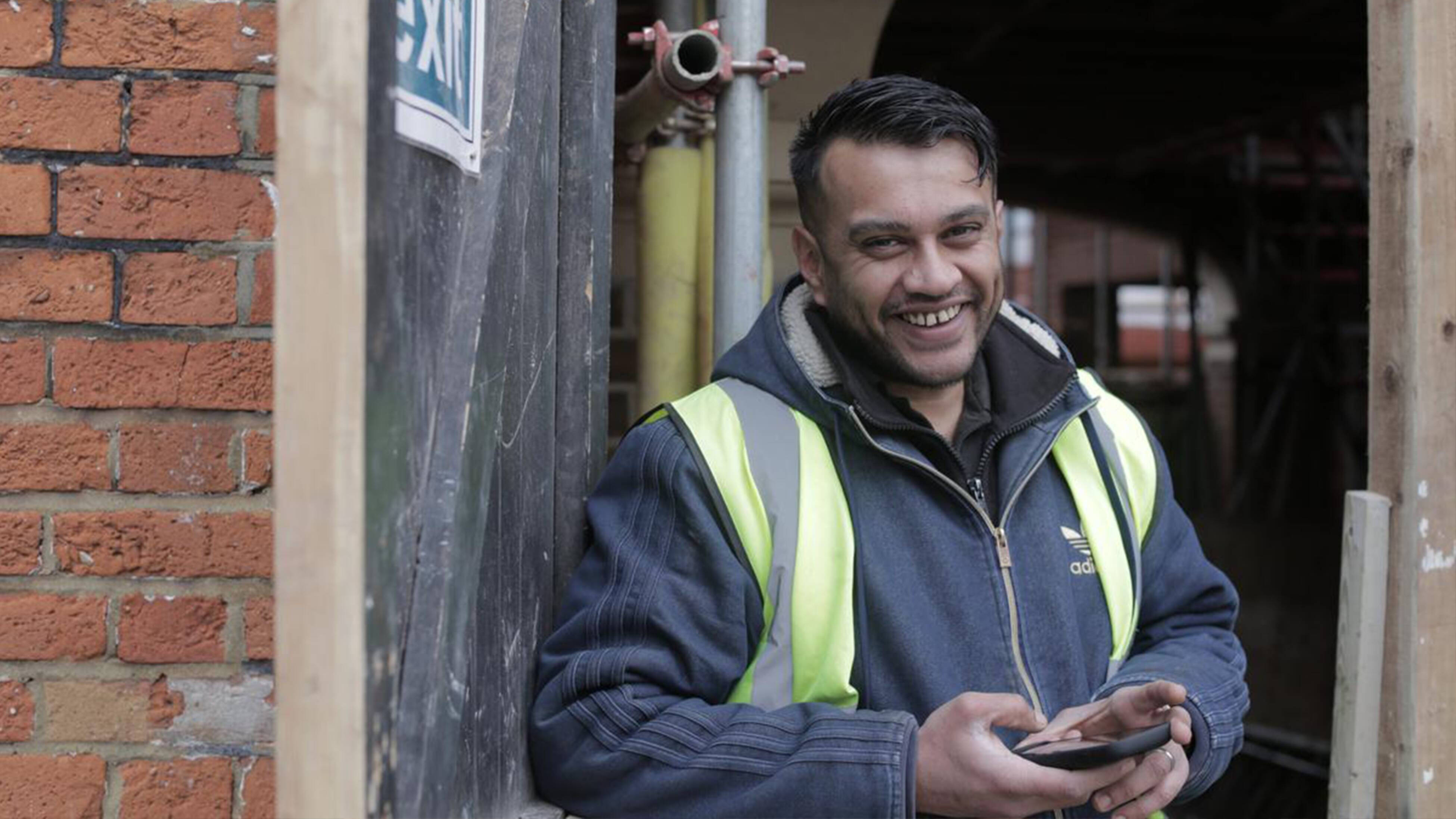 : A tradie wearing a high-visibility vest and jacket stands outdoors near a brick wall and scaffolding, holding a mobile phone