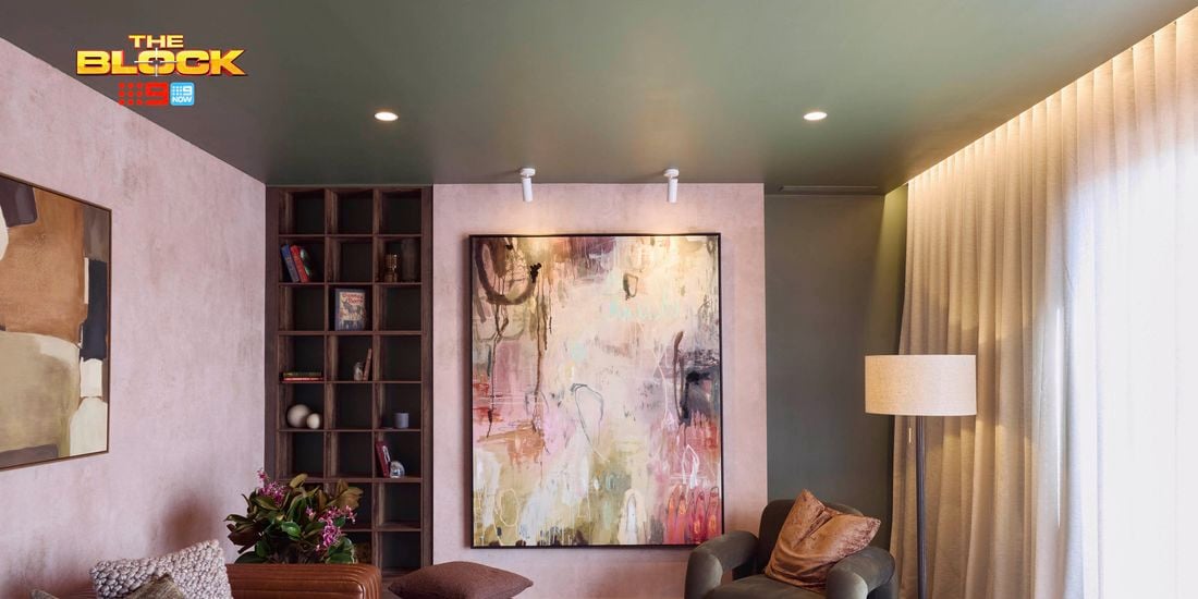 A modern living room featuring a large abstract painting on the wall, a brown leather sofa with cushions, an armchair, a wooden side table, and a floor lamp. A bookshelf with books and decorative items stands nearby. The room has soft lighting and curtains covering the windows. 