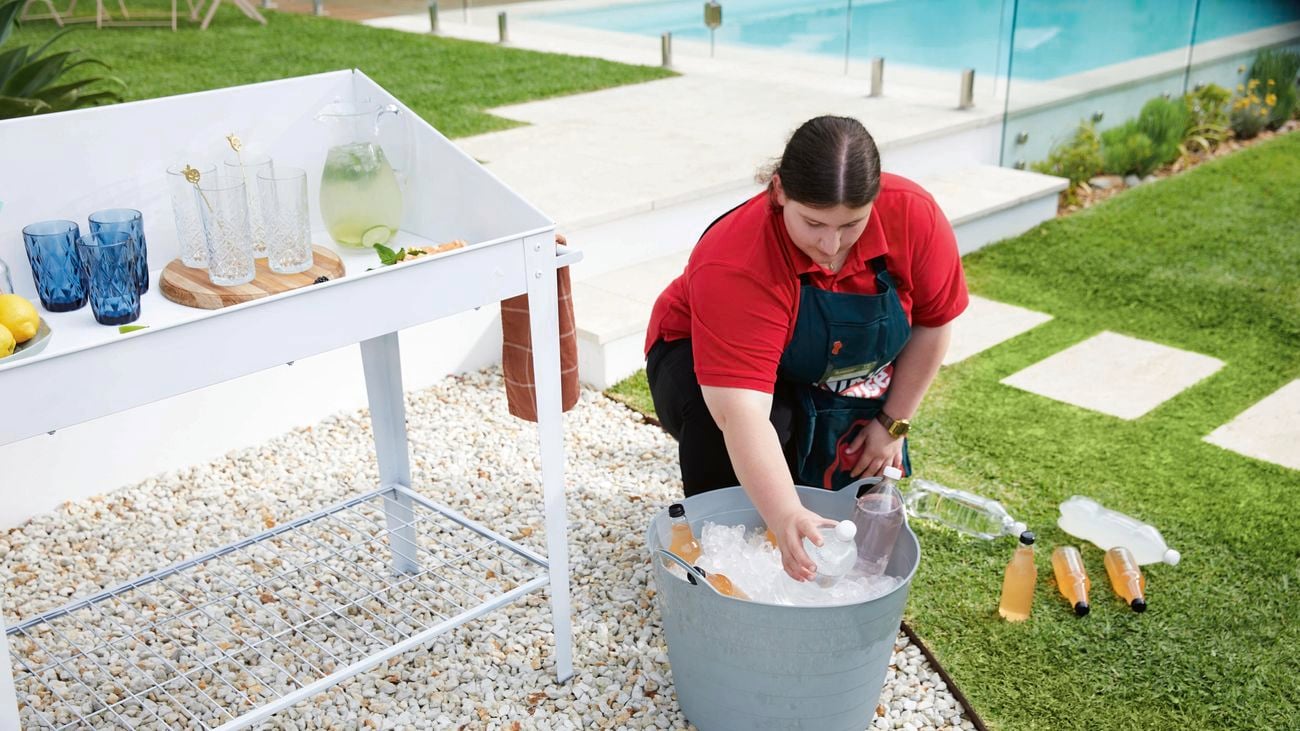 A Bunnings employee puts bottles in a bucket of ice next to a white drinks trolley with glasses and jug of punch on top.