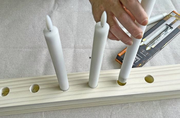  A hand inserts white flameless taper candles into drilled holes in a white wooden block, candle packaging is on the table.