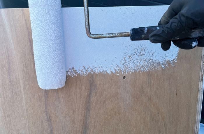 A gloved hand uses a roller to apply white paint to a timber surface.