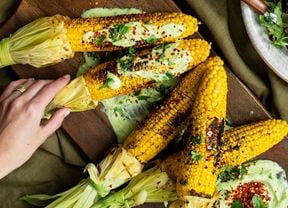 A wooden serving board with BBQ corn on the cob sitting on avocado sauce and topped with lime butter, chill and coriander