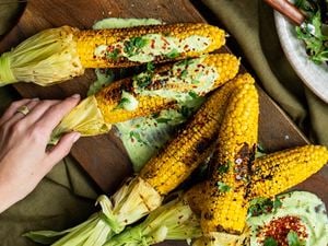 A wooden serving board with BBQ corn on the cob sitting on avocado sauce and topped with lime butter, chill and coriander