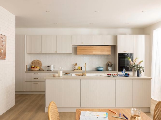 A beige modern kitchen, with a brown benchtop with crockery on it, an oven in it and a table and chairs in front of it.