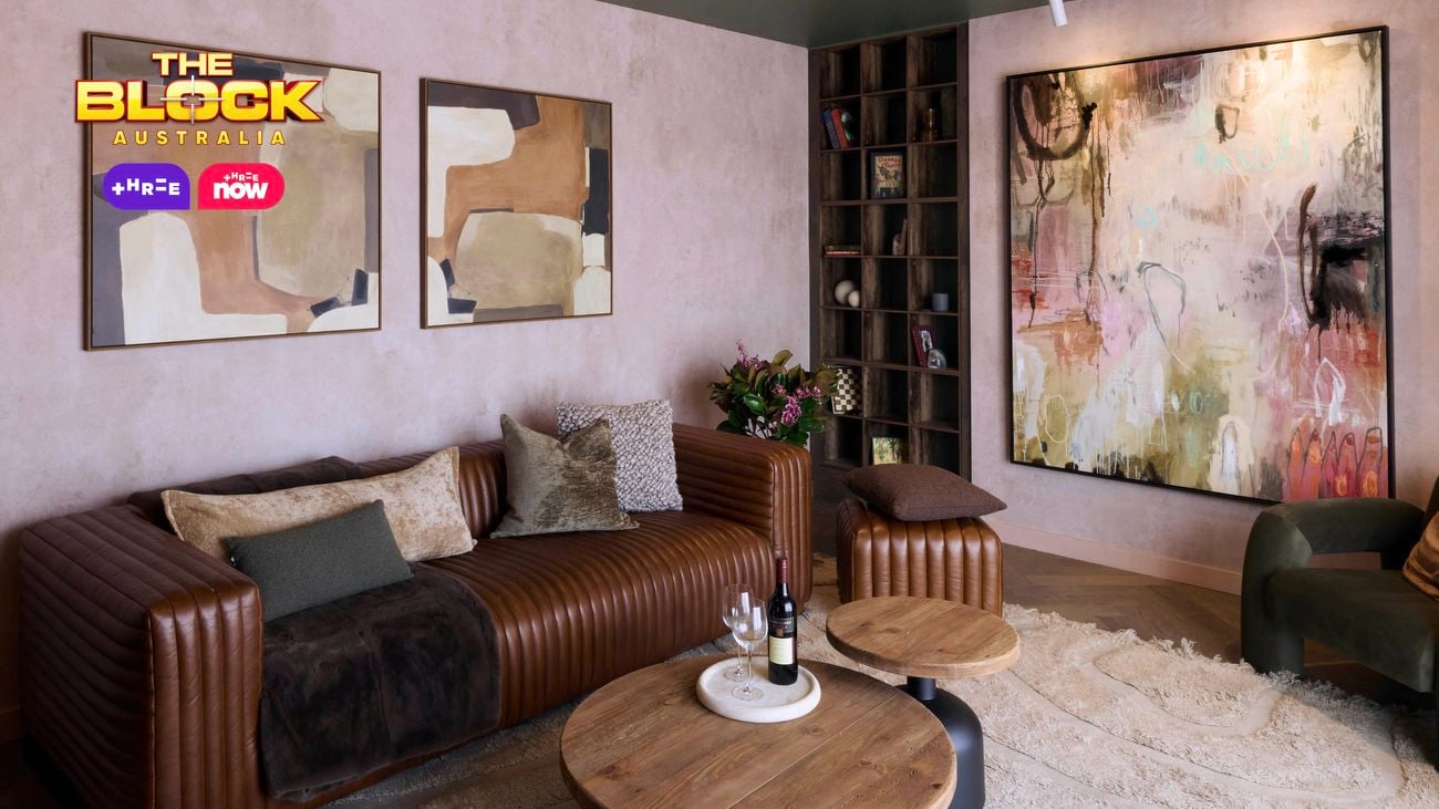 A modern living room featuring a brown leather sofa with assorted cushions, two round wooden coffee tables, and an abstract painting on the wall. Two smaller abstract artworks hang above the sofa. A bookshelf is in the corner, and a green armchair sits to the right. 
