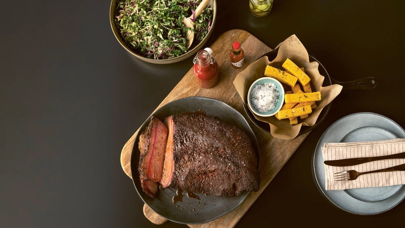 A sliced beef brisket on a wooden board, served with a side of coleslaw and polenta chips