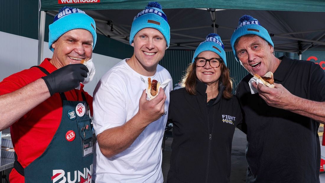 Four people wearing Biz Freeze blue beanies standing together under a Bunnings marquee, smiling while holding sausages in bread outdoors.