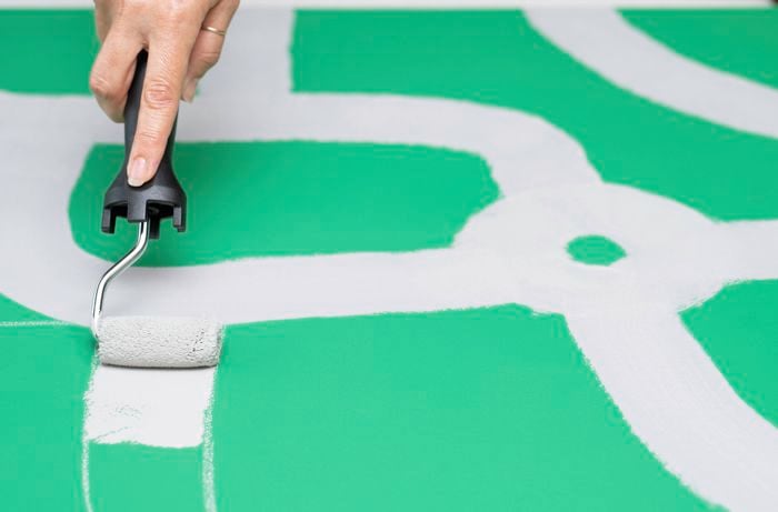 A person is applying grey paint on the roads along the pencil lines with a narrow roller.