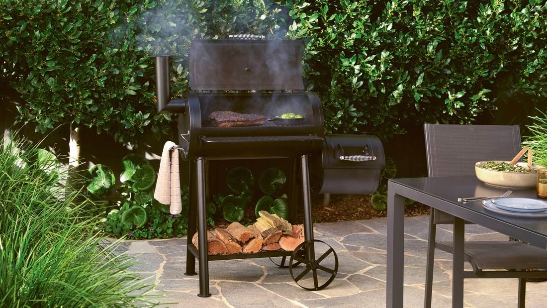  A black offset smoker barbecue with beef brisket and vegetables cooking inside, with firewood stored underneath.