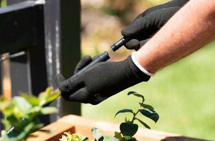 A gloved person connects two pieces of irrigation tubing using a plastic fitting in a garden setting.