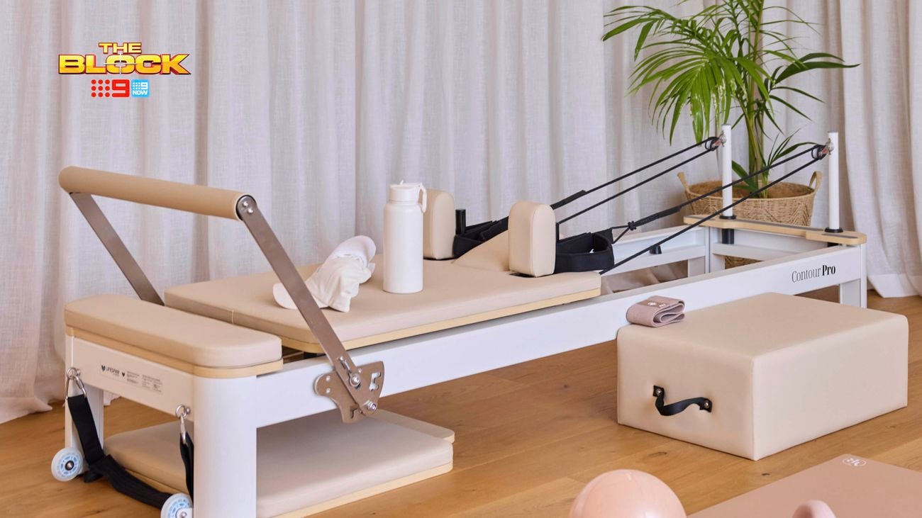 A Pilates reformer machine in a well-lit room, featuring a beige and white color scheme with padded sections. The machine is equipped with accessories including a water bottle, towel, and small fitness items. In the background, there’s a potted plant, an exercise ball, and foam rollers. 